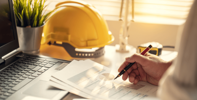 A person sitting at a desk writing on documents. A hard hat, laptop, and measuring tape are placed on the desk.