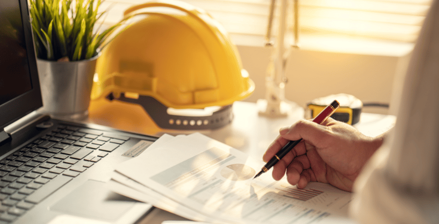 A person sitting at a desk writing on documents. A hard hat, laptop, and measuring tape are placed on the desk.