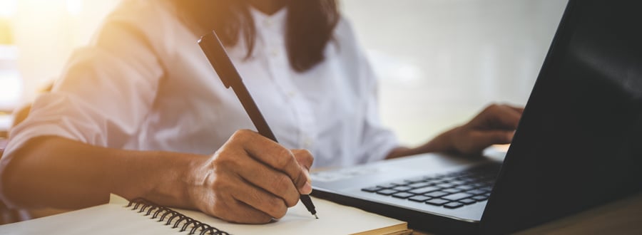 Woman working on laptop while taking notes on paper