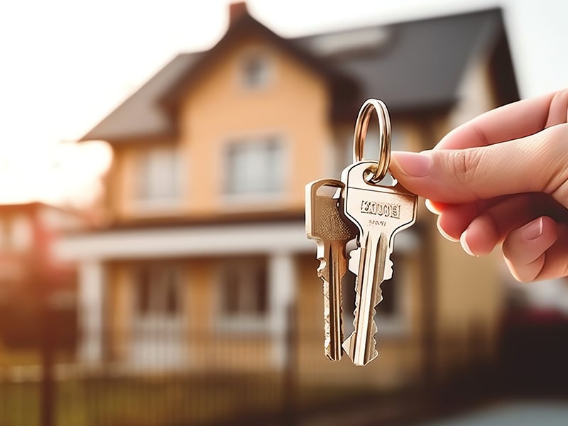 Closeup of a hand holding a set of keys to a house visible in the background